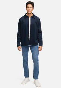 Mørkeblå skjorte med knapper og to forlommer, hvid T-shirt under, parret med lyseblå jeans og hvide sneakers. Glat stof.