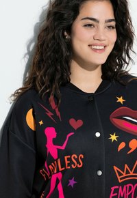 Schwarze Bomberjacke mit lebendigen grafischen Illustrationen in Pink, Orange und Gelb, mit lockerem Schnitt und Druckknopfverschluss.