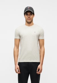 Άνδρας που φοράει ανοιχτό μπεζ κοντομάνικη μπλούζα και σκούρο παντελόνι, με μαύρο καπέλο που φέρει ένα μικρό κόκκινο, λευκό και μπλε λογότυπο.