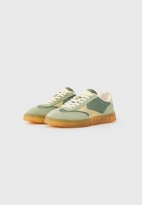 Grønne og beige sneakers med et teksturert stoffoverdel, gummisåle og kontrasterende snøringer. Har en særegen gjennomsiktig gummisåle.