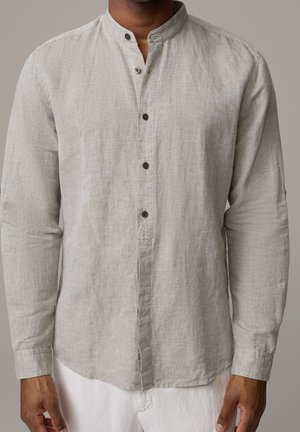 Uomo che indossa una camicia di lino grigio chiaro a maniche lunghe con collo alla coreana e bottoni neri, abbinata a pantaloni bianchi.