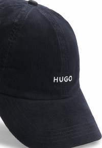 Sort corduroy-cap med en buet skygge og hvid broderet "HUGO" tekst på forsiden. Tekstureret stof med synlig ribbing.