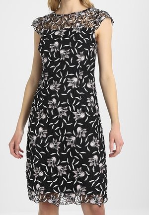 Frau trägt ein schwarzes ärmelloses Kleid mit weißen Blumen- und Blattmustern sowie einem transparenten Spitzenausschnitt und Saum.