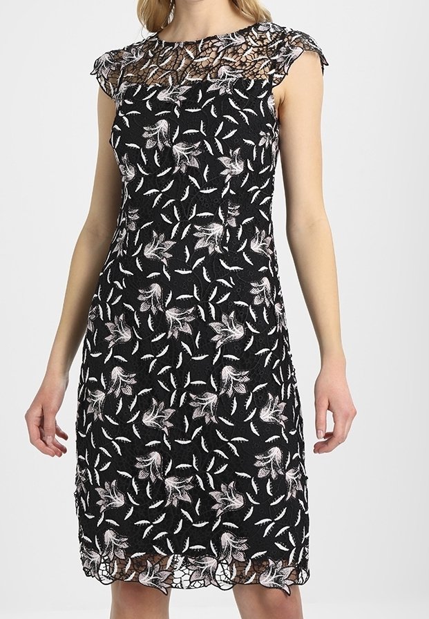 Femme portant une robe noire sans manches avec des motifs floraux et de feuilles blancs, avec un décolleté et un ourlet en dentelle transparente.