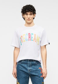 Άσπρο κοντομάνικο μπλουζάκι με πολύχρωμα γράμματα που σχηματίζουν τη λέξη "ICECREAM." Συνδυάζεται με μπλε τζιν τζινάκια με μπαλώματα.