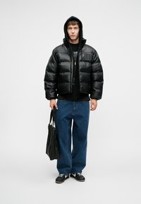 Sort dunjakke med blank finish, cropped design og logo detaljer. Kombineret med sort hoodie, løse blå jeans og sorte sko.