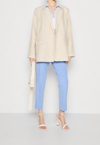 Beige double-breasted blazer with pocket details, light blue trousers, and light-colored strappy heels, holding a beige shoulder bag.