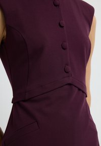 Robe sans manches en tissu bordeaux profond, présentant un design ajusté, une rangée de boutons ronds et un détail de couture avec une poche latérale cachée.