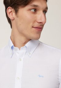 Chemise blanche à boutonnières avec une surface texturée, arborant un logo de teckel brodé bleu clair sur le côté gauche de la poitrine.