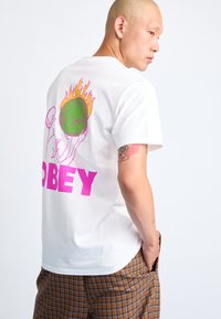 White cotton t-shirt featuring a bold pink "OBEY" design and colorful graphics of hands and a globe with flames on the back.
