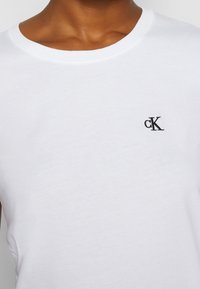 Vit bomullt-shirt med rund hals, korta ärmar och liten svart broderad logotyp på bröstet. Slät textur, klassisk passform.