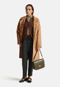 Hnědý vlněný kabát s kostkovaným vzorem, hnědý krácený kardigan přes šedé kalhoty, černé boty a zelenou texturovanou kabelku.