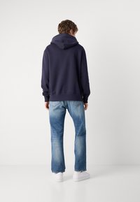 Mørkeblå hettegenser med stor hette, ribbestrikkede mansjetter og hem; parret med lyseblå jeans med rett ben og hvite sneakers. Ensfarget bakgrunn.
