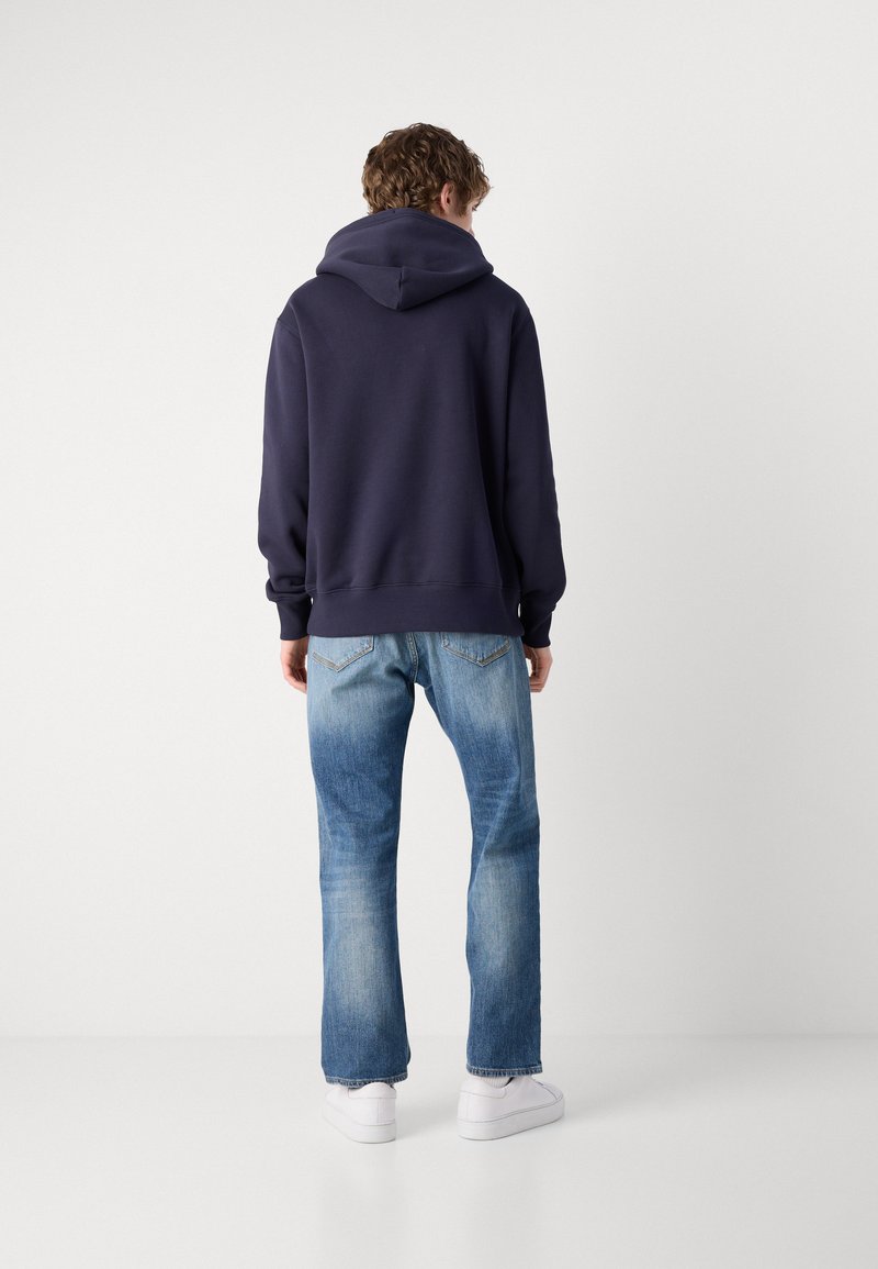 Felpa blu navy con un ampio cappuccio, polsini e fondo a costine; abbinata a jeans dritti azzurri e sneaker bianche. Sfondo semplice.