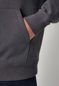 Sweatshirt gris foncé avec des poignets et un ourlet côtelés, doté d'une poche latérale. Le matériau a une texture lisse avec une finition légèrement brossée.