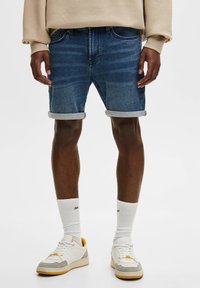 Shorts en jean bleu foncé avec un effet délavé clair, revers roulés, cinq poches, associés à des baskets blanches et des chaussettes blanches.