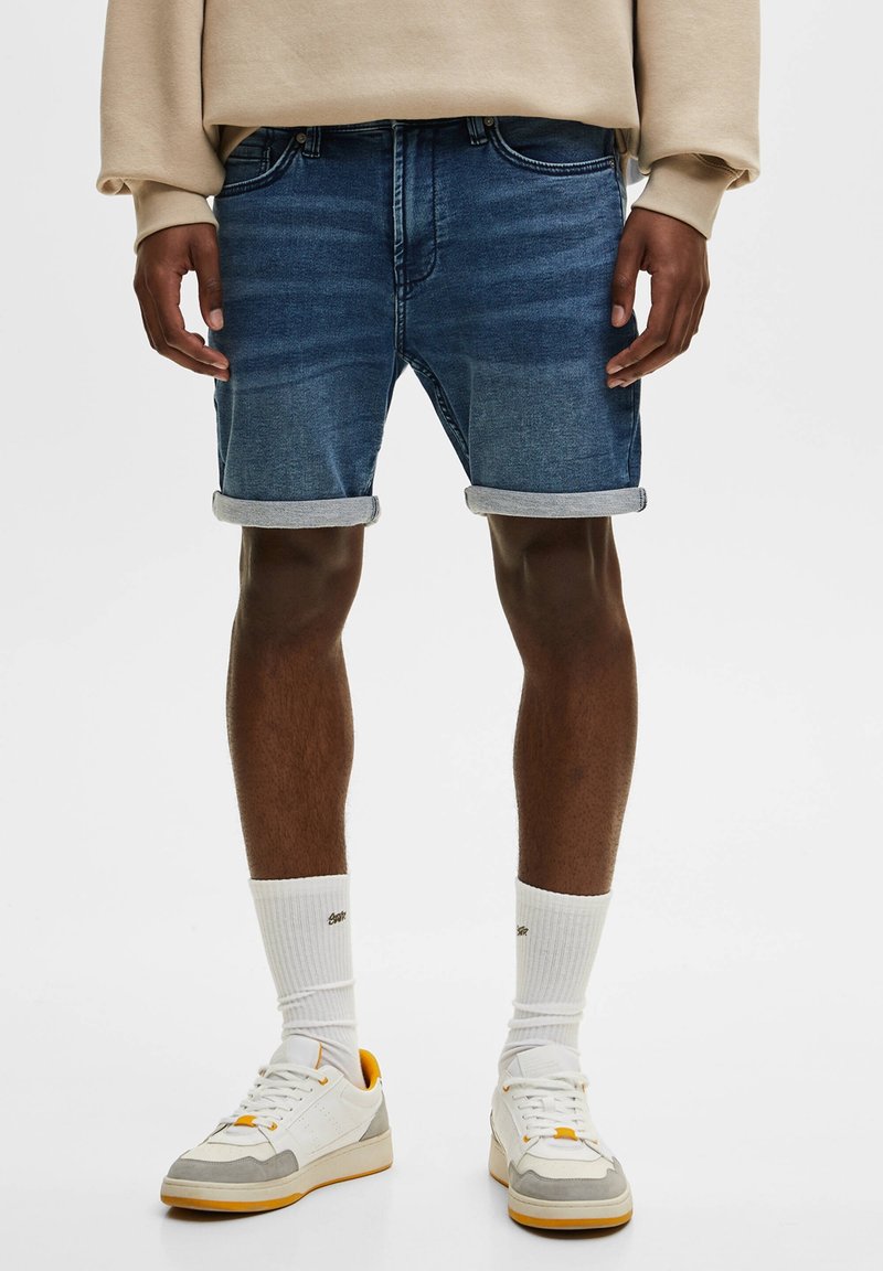 Shorts en jean bleu foncé avec un effet délavé clair, revers roulés, cinq poches, associés à des baskets blanches et des chaussettes blanches.