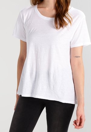 Vrouw draagt een effen wit T-shirt met korte mouwen en zwarte broek, met een kleine 'Helene' tatoeage op haar linker onderarm.