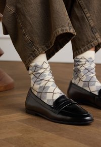 Black leather loafers with a smooth finish, styled with cream and gray argyle patterned socks, paired with loose brown pants.