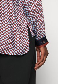 Long-sleeve shirt in light blue fabric, featuring red polka dots. Hem is split with navy contrast cuffs and a brown button detail.