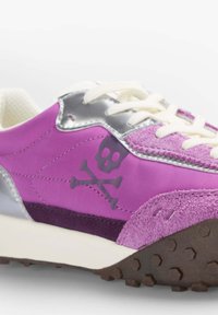 Baskets violettes présentant des textures lisses et en daim, des accents argentés, des lacets crème, et un logo tête de mort et os croisés sur le côté.