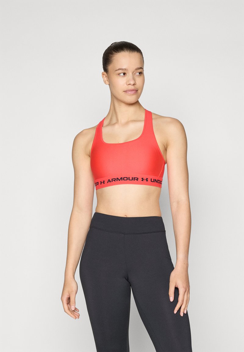 Rød sports-bh med firkantet halsutskjæring, laget av glatt materiale, med et svart logobånd nederst. Kombinert med svarte leggings.