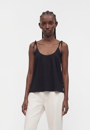 Femme aux cheveux tressés portant un top noir sans manches avec des bretelles à nouer et un pantalon blanc, debout devant un fond clair uni.