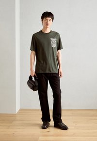Olivengrøn t-shirt med grafisk print på venstre bryst, parret med sorte lige bukser og sorte slip-on sko. Holder en sort håndtaske.