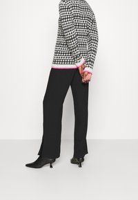 Schwarzer Strickpullover mit weißem Muster und rosa Besatz, kombiniert mit schwarzen Hosen mit Seitenschlitz und spitzen schwarzen Absatzstiefeln.