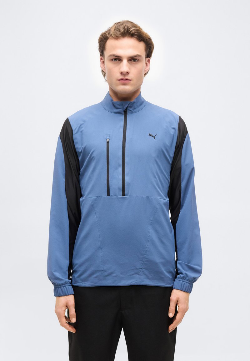 Νεαρός άνδρας που φοράει μπλε και μαύρο μπουφάν αθλητικής φόρμας Puma με ψηλό γιακά και τσέπη με φερμουάρ, στέκεται μπροστά σε απλό ανοιχτό φόντο.