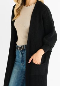 Černý pletený cardigan s otevřeným předním dílem, oversized rukávy a bočními kapsami. Nositelný přes béžový vypasovaný top a světle modré džíny.