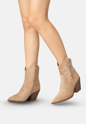 Jambes portant des bottes de cowboy en daim beige mi-mollet avec talons blocs, fermetures éclair latérales et surpiqûres décoratives sur un fond uni.