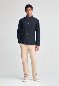 Dunkelblaues Hemd mit langen Ärmeln, das ein kleines rotes Logo zeigt. Kombiniert mit beigen Hosen und hellen Sneakers.