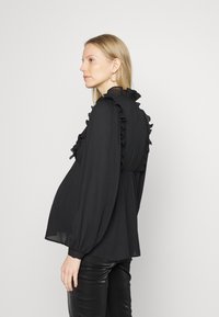 Blusa preta com detalhes em babados ao longo da gola e da parte frontal, mangas longas bufantes e uma silhueta solta. Apresenta uma textura de tecido transparente.