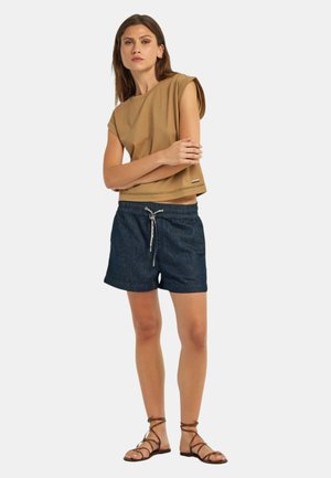 Donna con capelli castani che indossa una canotta beige, pantaloncini blu navy con coulisse e sandali marroni con cinturini, in piedi con le braccia incrociate.
