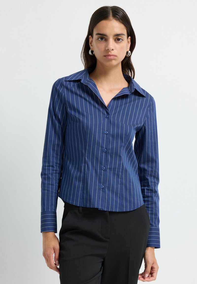 Camicia abbottonata blu navy con strisce verticali bianche, maniche lunghe, colletto a punta e orlo leggermente curvato, realizzata in un tessuto morbido.