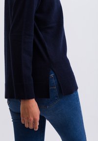 Námořnický vlněný svetr s žebrovanými manžetami a bočními rozparky, spárovaný s těsnými modrými džíny. Je patrná hladká textura a ležérní design.