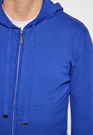 Blaue Zip-Hoodie mit einer Kordelzugkapuze, aus strukturiertem Strickstoff und mit Metallreißverschluss-Details. Nahaufnahme des oberen Abschnitts.