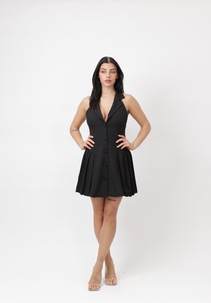 Frau mit langen dunklen Haaren, die ein ärmelloses schwarzes Kleid mit Knöpfen und plissiertem Rock trägt, steht mit den Händen in der Hüfte vor einem weißen Hintergrund.