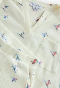 Oberteil aus cremefarbenem strukturiertem Stoff mit V-Ausschnitt, versehen mit bunten Skifahrer-Illustrationen und einer Tasche auf der Vorderseite.