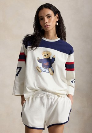 Femme aux longs cheveux bruns portant un sweat-shirt blanc et bleu marine oversize avec un motif d'ourson en peluche et un drapeau, associé à un short blanc à cordon de serrage.