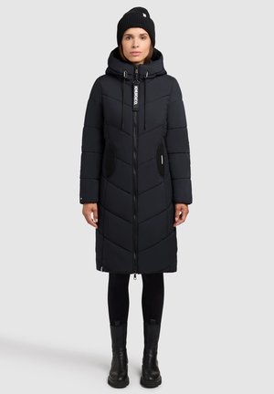 Femme portant un long manteau matelassé noir avec capuche, un bonnet en tricot noir, des leggings noirs et des bottines noires, se tenant devant un arrière-plan uni.