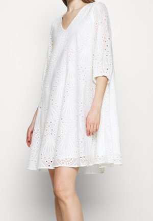 Woman wearing a white, knee-length eyelet dress with three-quarter puff sleeves and a V-neckline, standing against a plain background.