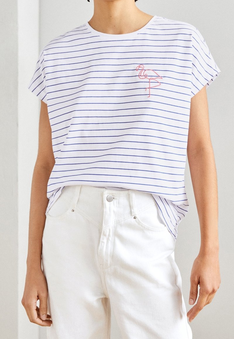 Camiseta blanca a rayas con rayas horizontales azules, con un flamenco rosa bordado en el pecho. Corte relajado, mangas cortas.