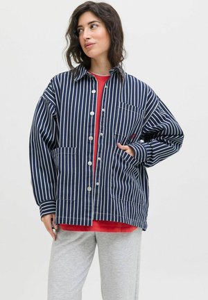Vrouw in een marineblauw en wit verticaal gestreept button-up jasje over een rode shirt, met lichtgrijze broek, staand tegen een effen achtergrond.