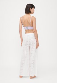 Lila bralette állítható pántokkal és virágos folttal, fehér, bő szárú nadrággal párosítva, amely szem alakú kivágásokkal és virágmintákkal rendelkezik.