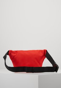 Rode stoffen heuptas met een zachte textuur, voorzien van een zwarte verstelbare nylonriem en een plastic gespsluiting. Eenvoudig ontwerp, zonder aanvullende patronen.