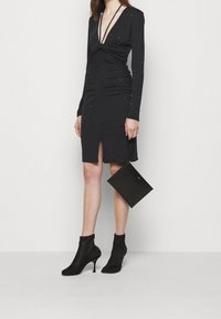 Robe noire à manches longues avec un décolleté en V profond, taille froncée, accents scintillants, fente haute jusqu'à la cuisse, portée avec des bottines noires et un petit sac à main.