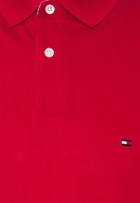 Rød polo-shirt lavet af struktureret stof, med en klassisk krave, tre hvide knapper og et lille broderet logo i blå, hvid og rød.