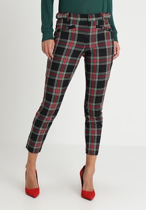 Žena oblečená v černých, červených a bílých károvaných upnutých kalhotách s zeleným svetrem a červenými lodičkami stojí na bílém pozadí.